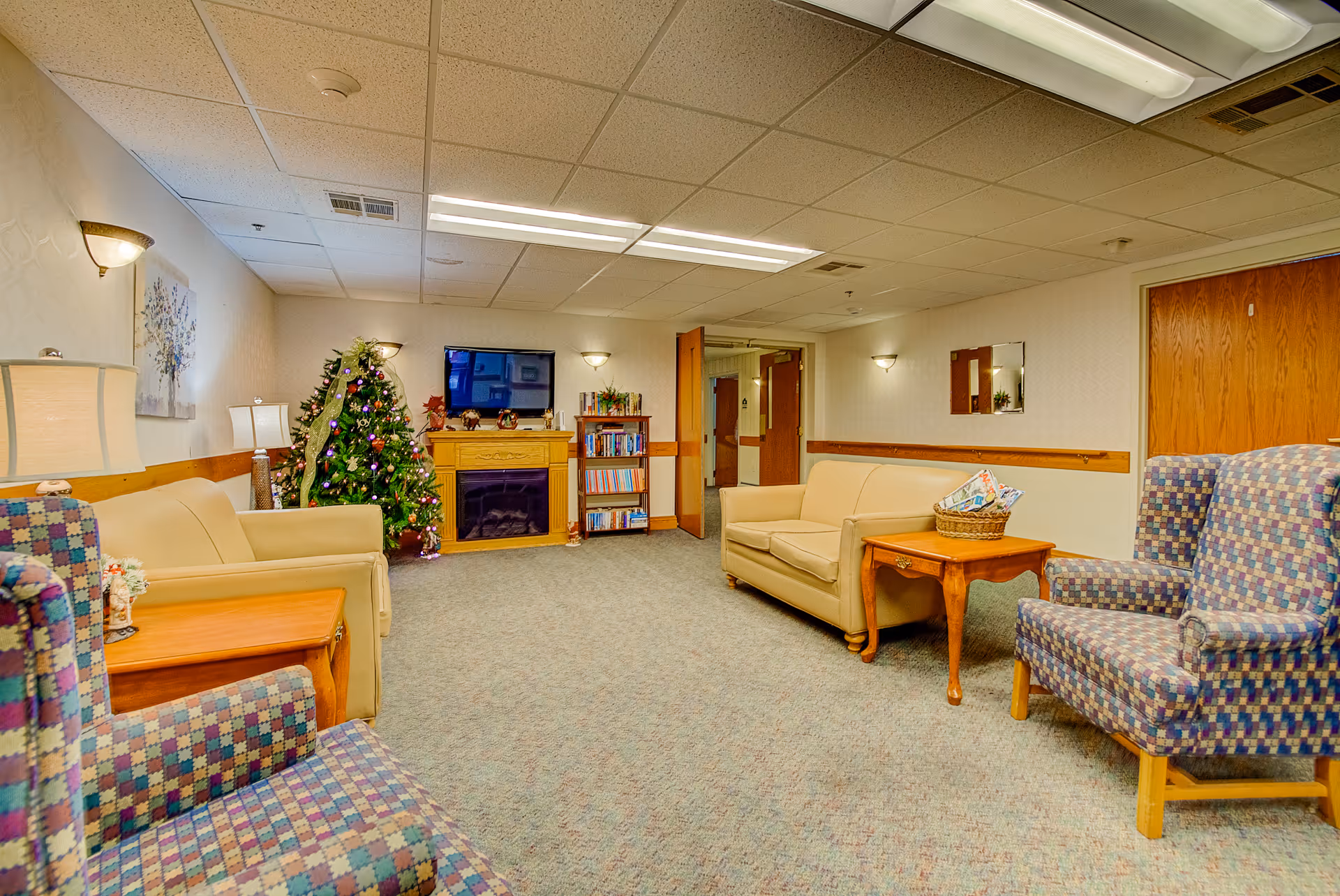 A cozy living room area in a senior living facility with beige sofas, patterned armchairs, a wooden side table with a basket of magazines, a decorated Christmas tree, a bookshelf, a wall-mounted TV above a wooden electric fireplace, and soft lighting from wall sconces and lamps.