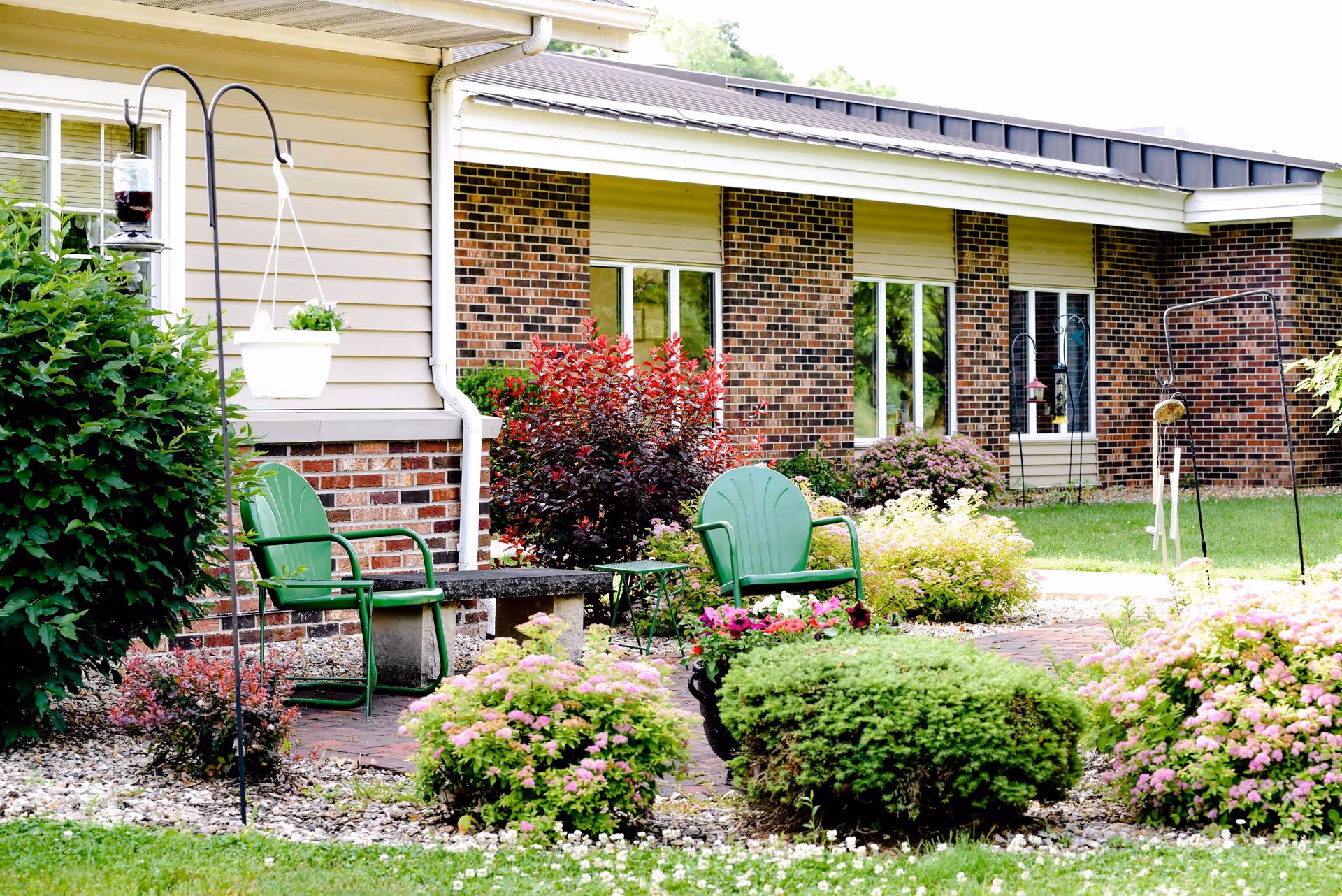 Outdoor garden area at Maple Crest Campus featuring green metal chairs, a stone bench, hanging flower pots, various flowering bushes, and a brick building with windows in the background.
