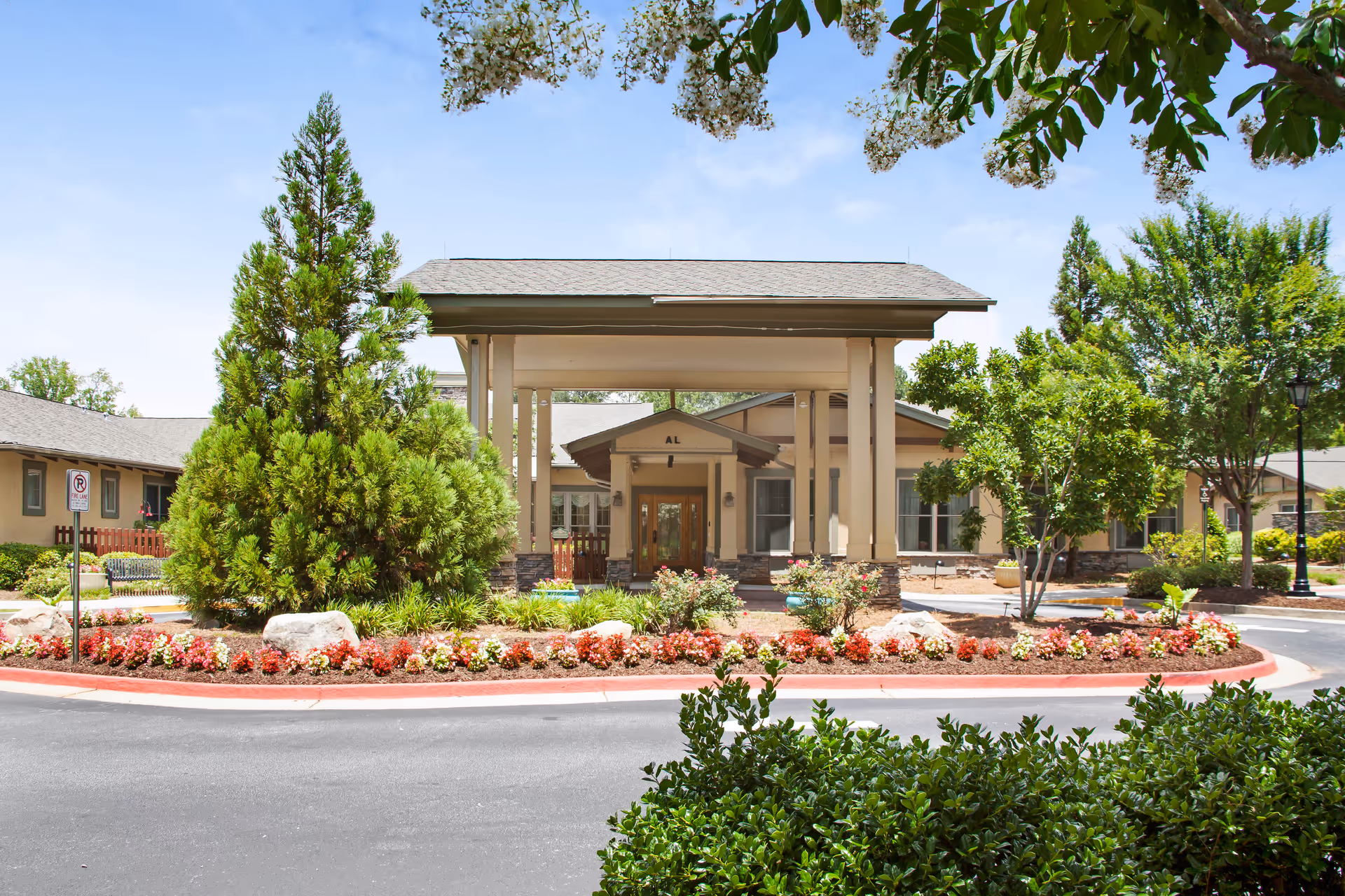 Front entrance of a senior living facility with a covered porte-cochère, circular landscaped drive, and flowering beds.