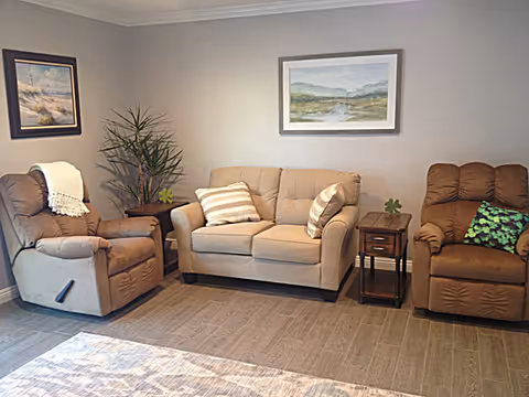 A cozy living room with a beige loveseat flanked by two brown recliner chairs. The loveseat has two striped throw pillows. There are two small wooden side tables with plants on them, and two framed landscape paintings hang on the light gray walls. The floor is covered with wood-like tiles and a patterned area rug.