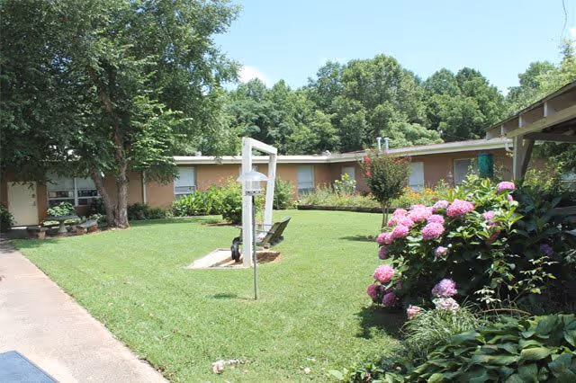 Courtyard with a green lawn, pink hydrangeas, benches, and a single-story brick building surrounded by trees.