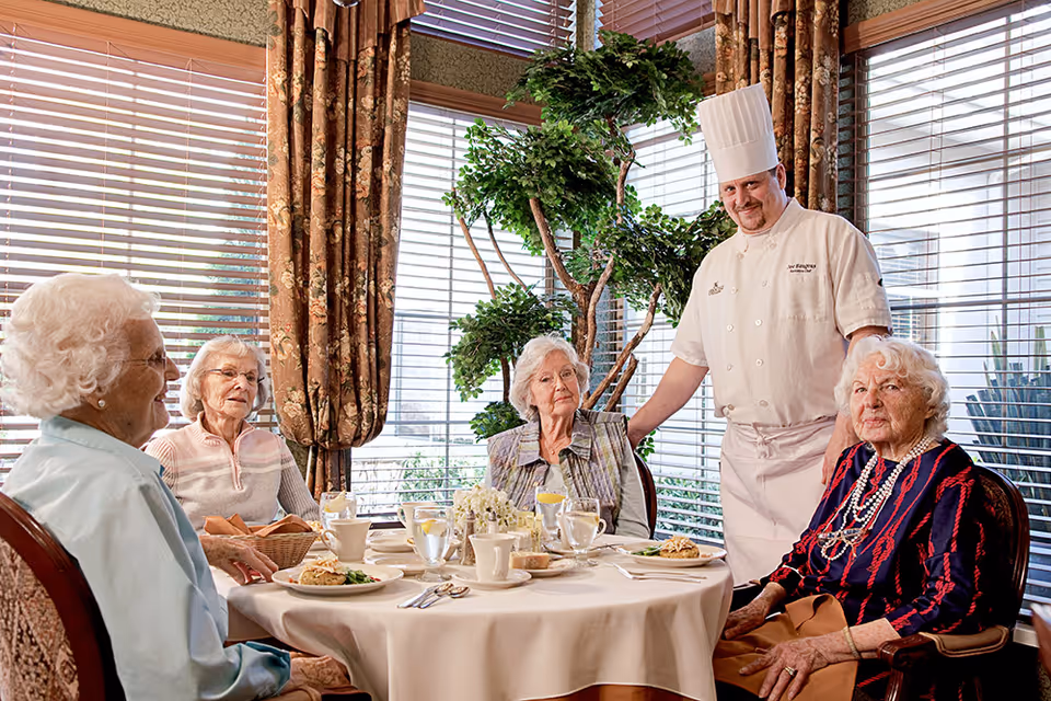 A chef standing beside a dining table with four elderly women seated around it, enjoying a meal in a well-lit room with large windows and floral curtains.