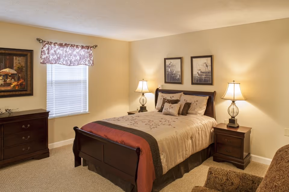 A cozy bedroom with a wooden bed frame, beige and brown bedding, two bedside tables with lamps, a wooden dresser, a window with a patterned valance, two framed bird pictures on the wall, and a patterned armchair in the corner.