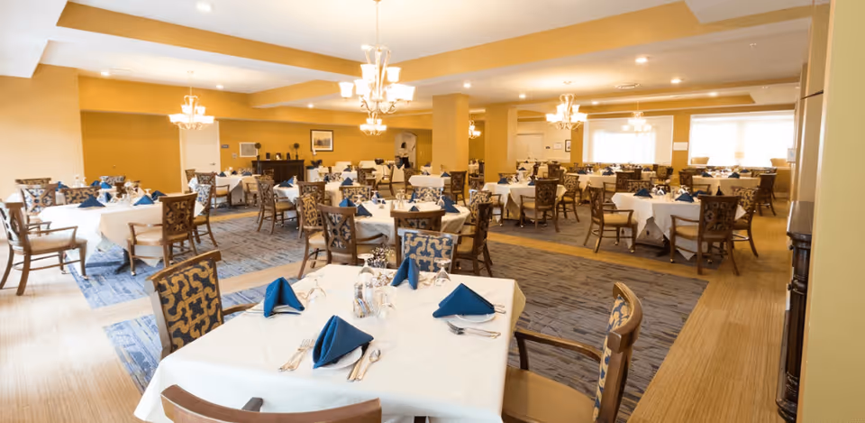 A spacious dining room with multiple round tables covered in white tablecloths, each set with blue folded napkins, silverware, and glassware. The room features patterned chairs, warm yellow walls, carpeted and wooden flooring sections, and elegant chandeliers hanging from the ceiling.