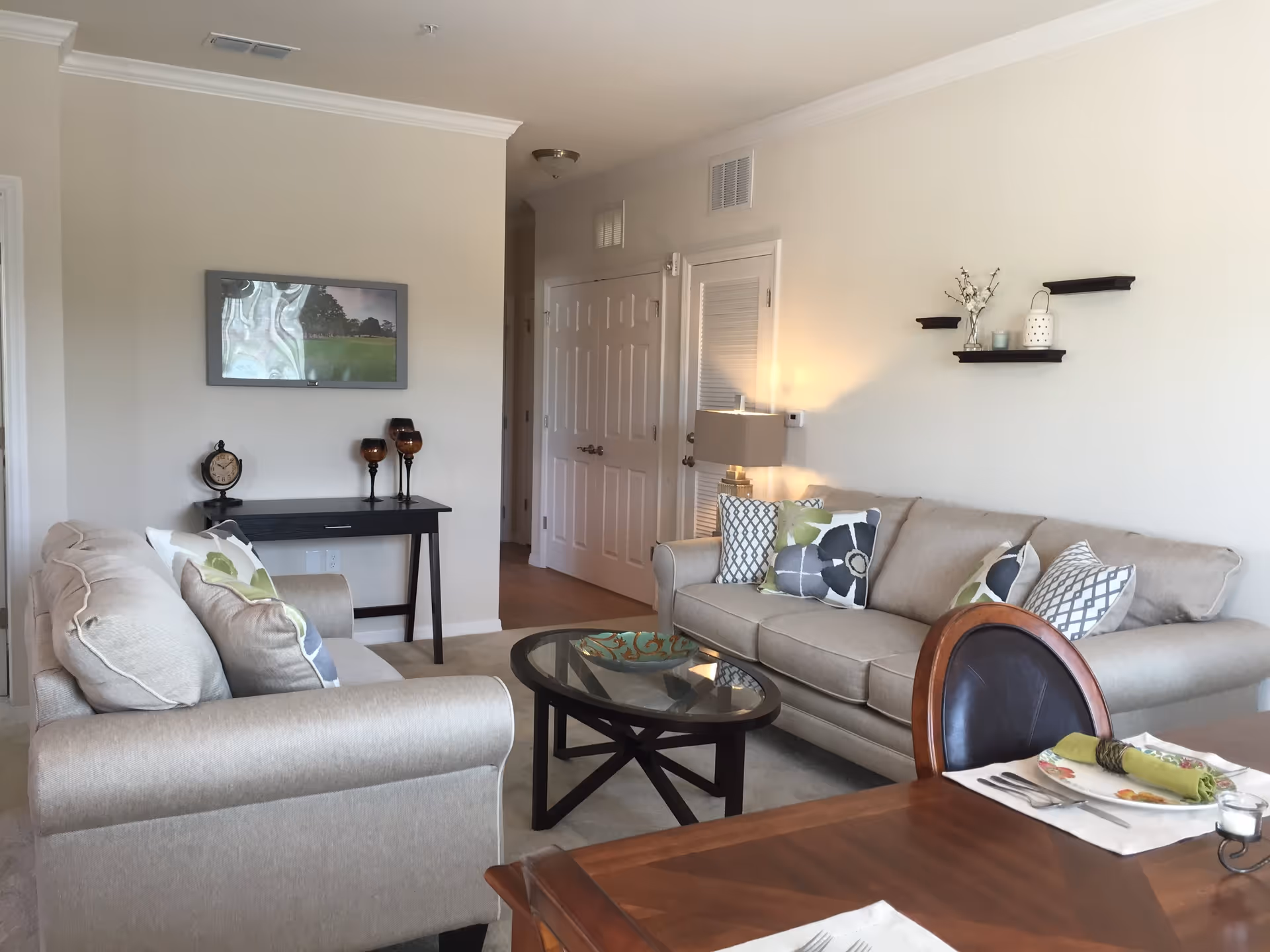 A bright living room with two beige sofas adorned with patterned cushions facing each other across a round glass coffee table. A small black console table with decorative items and a framed picture hangs on the wall behind one sofa. On the opposite wall, two small black shelves hold decorative items above one sofa. Part of a wooden dining table with a place setting is visible in the foreground.