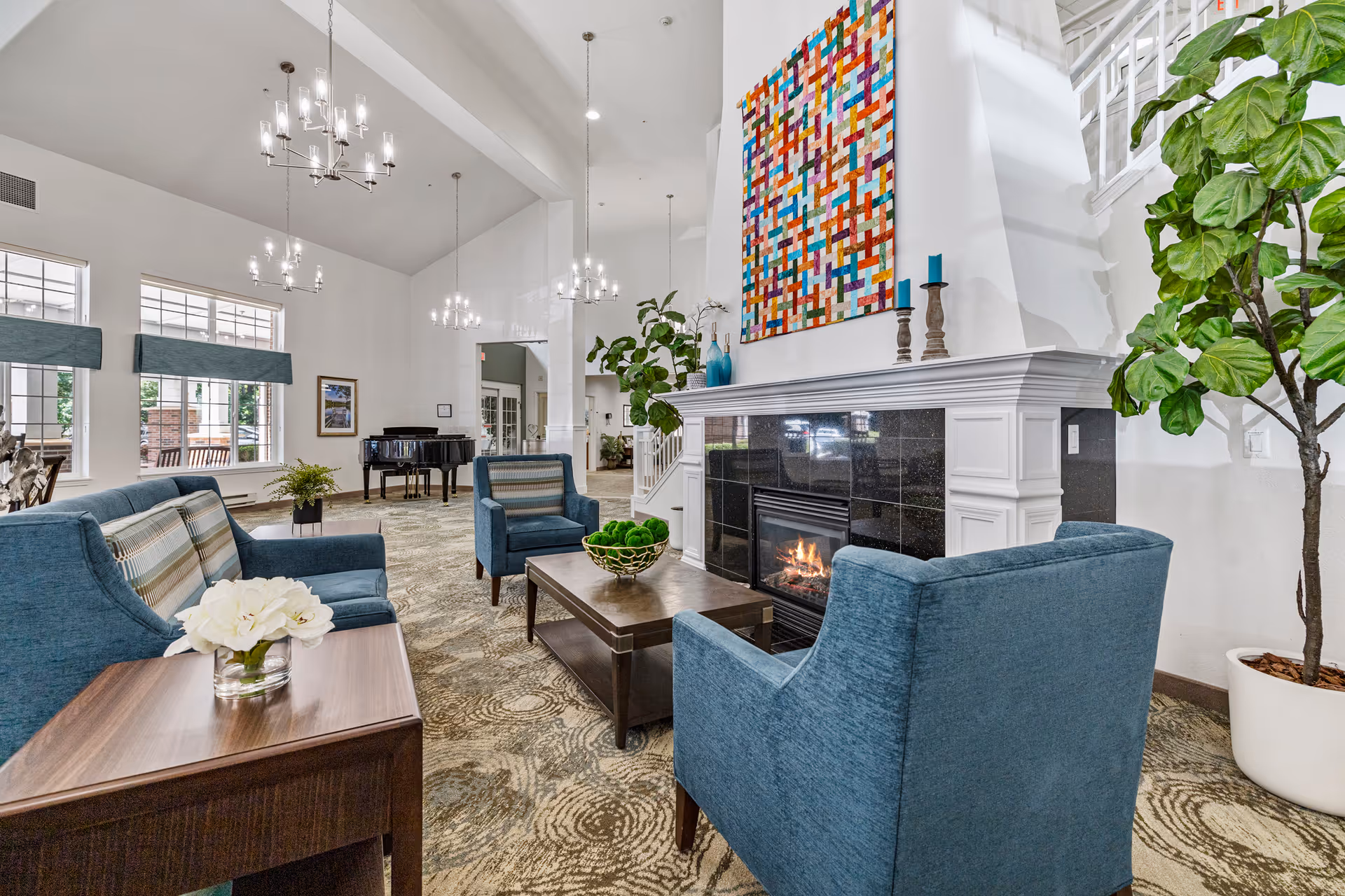 A spacious and well-lit living room area with high ceilings, featuring blue upholstered chairs and sofas arranged around a wooden coffee table with a bowl of green decorative balls. There is a fireplace with a black tile surround and a colorful abstract artwork hanging above it. Large windows with blue valances allow natural light to fill the room. A grand piano is visible in the background, along with several chandeliers hanging from the ceiling and potted plants adding greenery to the space.