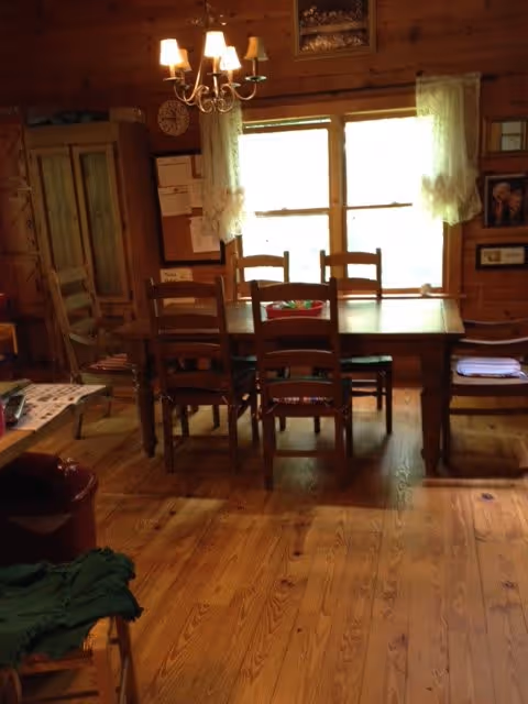 A cozy dining room with wooden floors, walls, and furniture. There is a rectangular wooden dining table surrounded by six wooden chairs. A chandelier with five lights hangs above the table. Two windows with sheer white curtains allow natural light into the room. Various framed pictures and a clock are mounted on the walls.