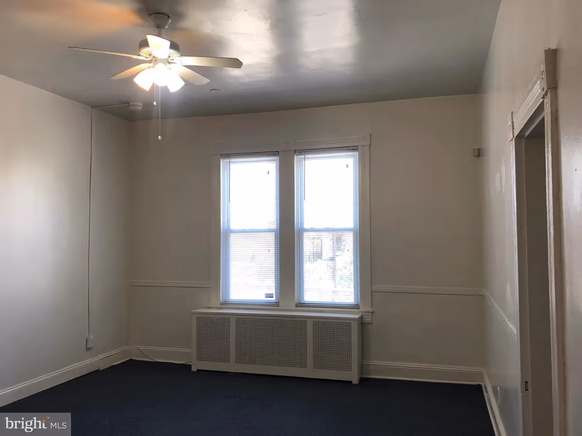 Empty room with white walls, a ceiling fan with lights, two windows with blinds, a radiator cover below the windows, and a doorway on the right side.