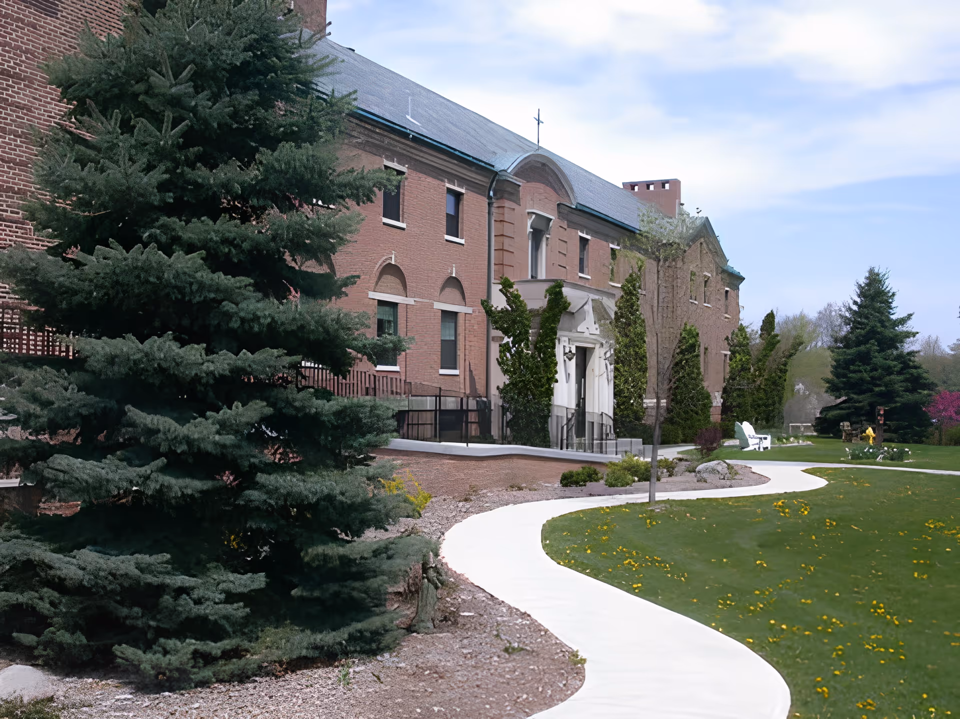 Front exterior of a brick building with a curved white walkway, large evergreen trees, and a grassy lawn.