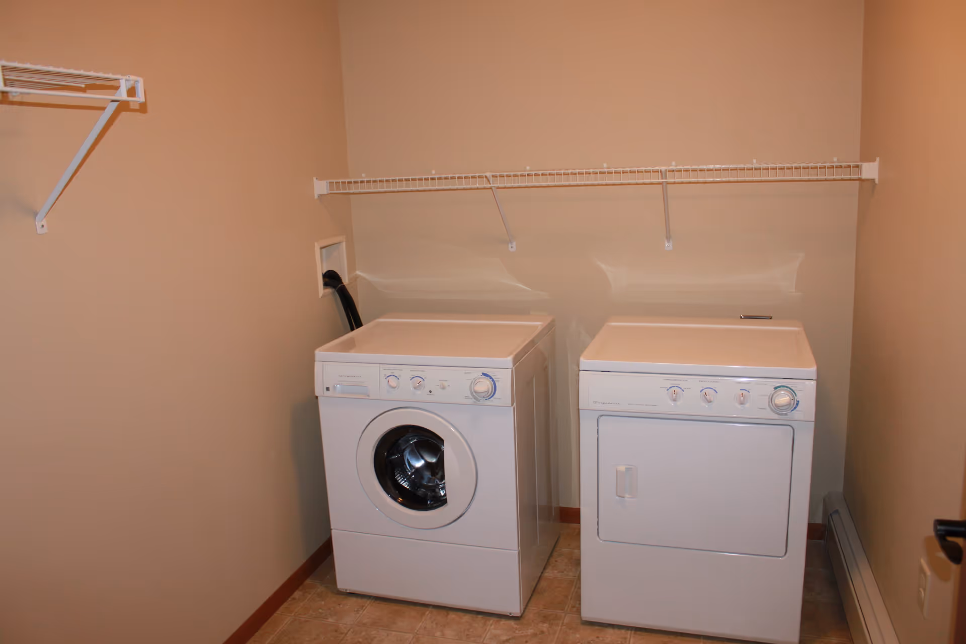 A laundry room with a white front-loading washing machine and a white dryer side by side against a beige wall. Above the machines is a white wire shelf mounted on the wall. The floor is tiled and there is a baseboard heater along the right wall.