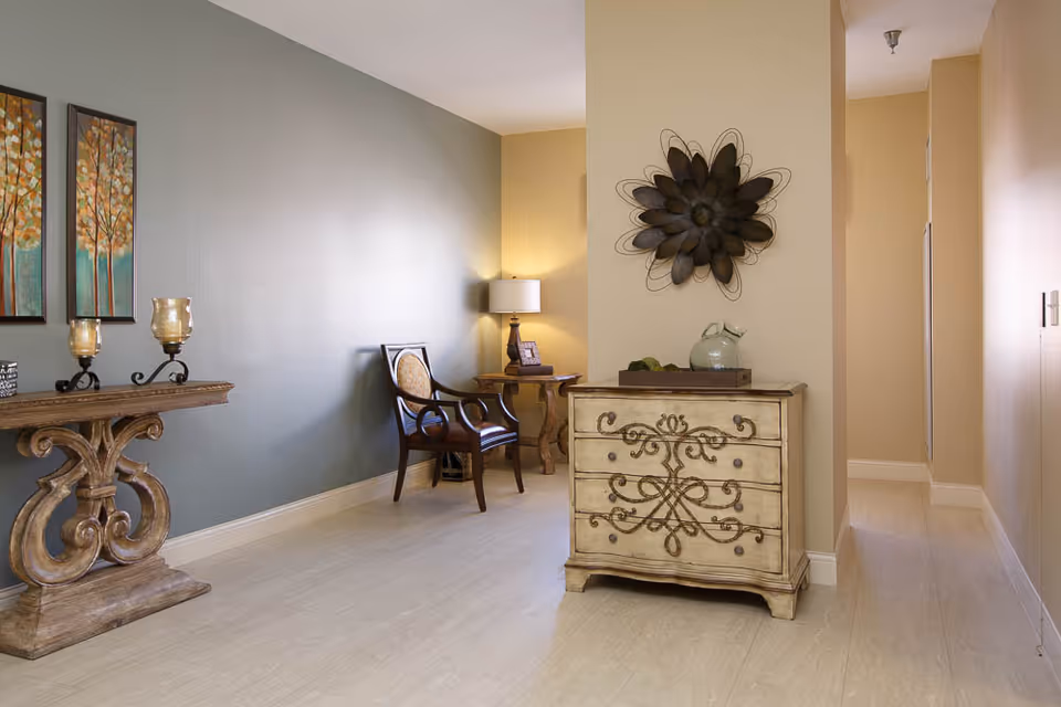 A well-lit interior hallway area with beige and light blue walls. There is a decorative wooden console table with two candle holders and two framed paintings of trees on the left side. A wooden chair with a cushioned seat and backrest is placed against the blue wall. Next to the chair is a small wooden side table with a lamp and a framed photo. In the foreground, there is an ornate wooden chest with intricate designs and a decorative metal flower wall art above it. The floor is light-colored tile, and a hallway extends to the right.