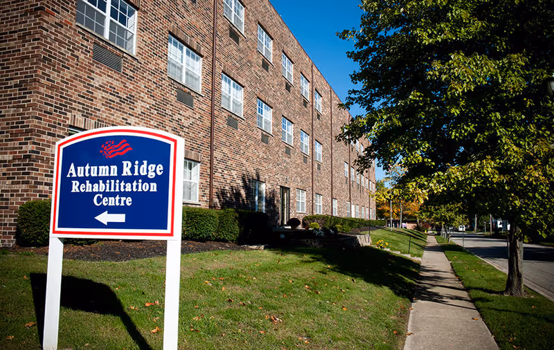 Exterior view of a brick building with multiple windows, a sidewalk, and a green lawn with trees. A blue and white sign in the foreground reads 'Autumn Ridge Rehabilitation Centre' with an arrow pointing left.