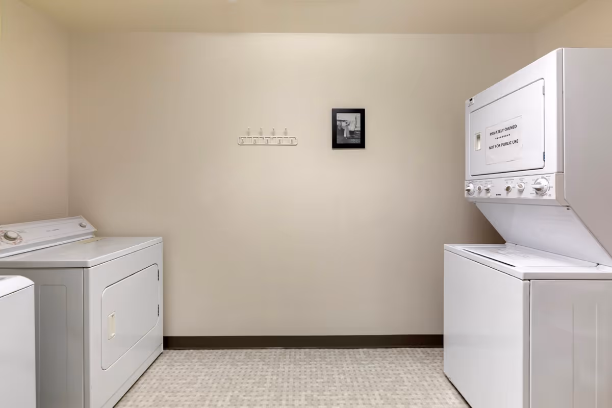 Laundry room with a white stacked washer and dryer on the right and a separate white dryer on the left. The walls are beige with a small black and white framed picture and a white wall-mounted rack with hooks in the center.