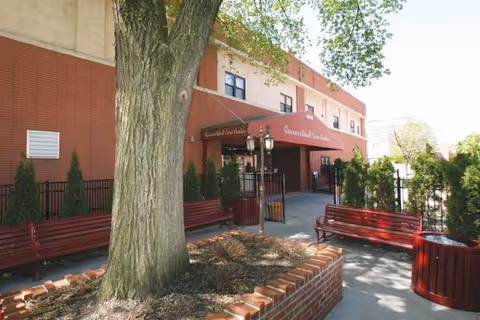 Exterior view of Queens Adult Care Center with benches, trees, and brick planters.
