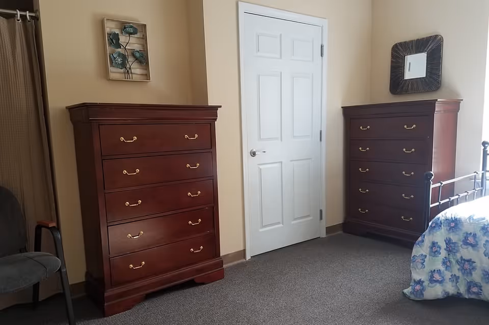 Bedroom with two tall wooden dressers flanking a white door, a partial iron bed with floral bedding, and a chair.