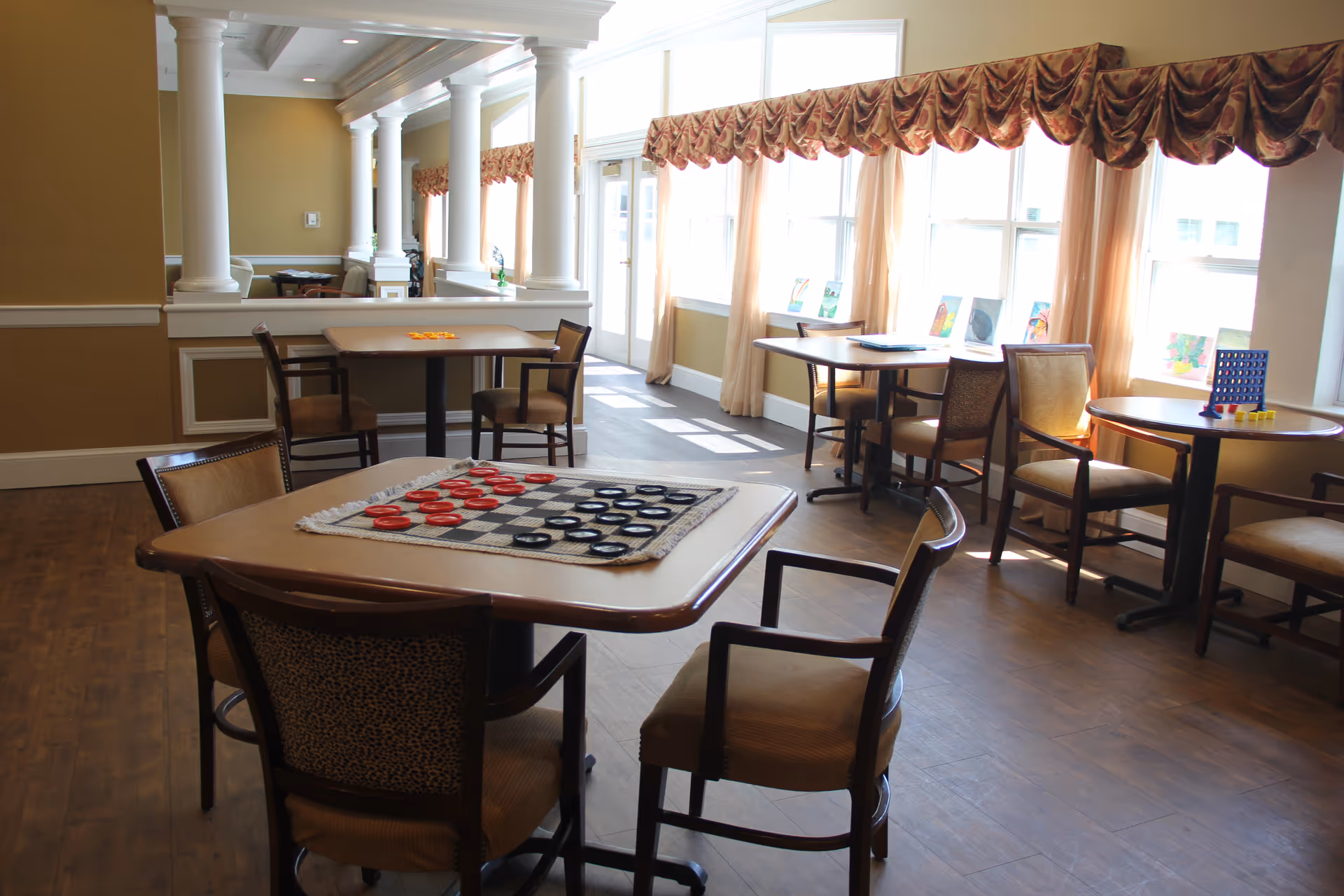 A bright and spacious common area with several tables and chairs arranged for social activities. One table in the foreground has a checkers game set up, while another table has a Connect Four game. Large windows with decorative valances allow natural light to fill the room, and there are columns and beige walls in the background.