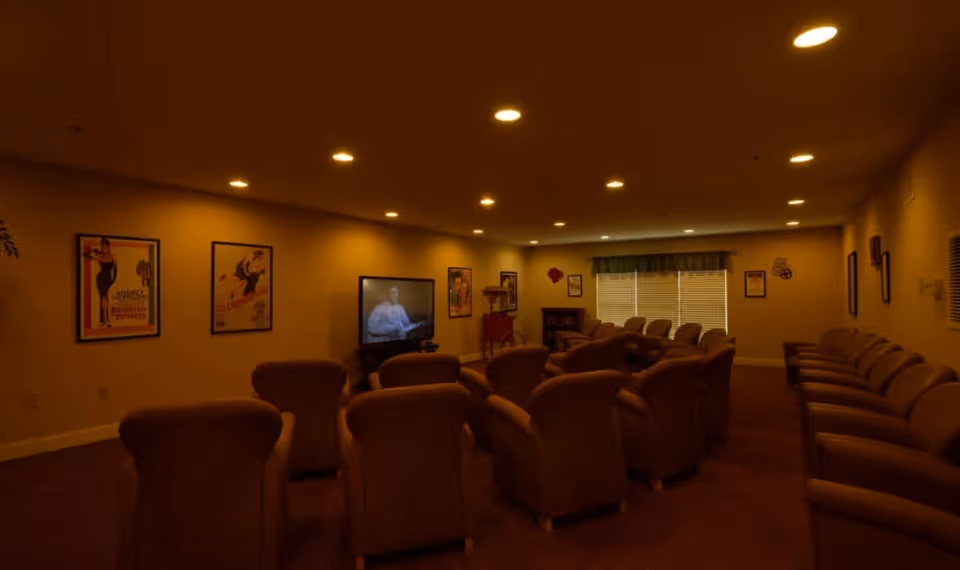A dimly lit common room with multiple rows of cushioned chairs facing a television screen mounted on the wall. The walls are decorated with framed vintage posters, and there is a window with blinds and a valance at the far end of the room.