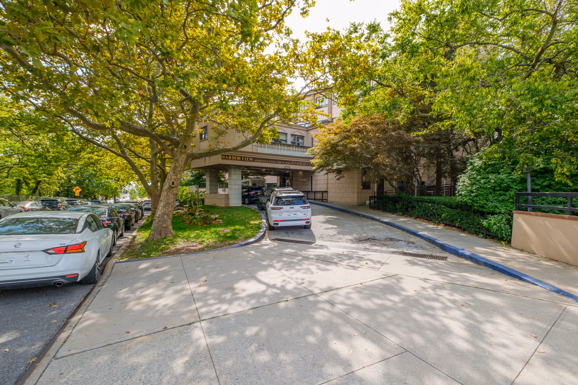 Entrance driveway of Harbor View Home, an assisted living facility, with a covered drop-off area. Several cars are parked along the street and in the driveway. The building is surrounded by large green trees and landscaping.