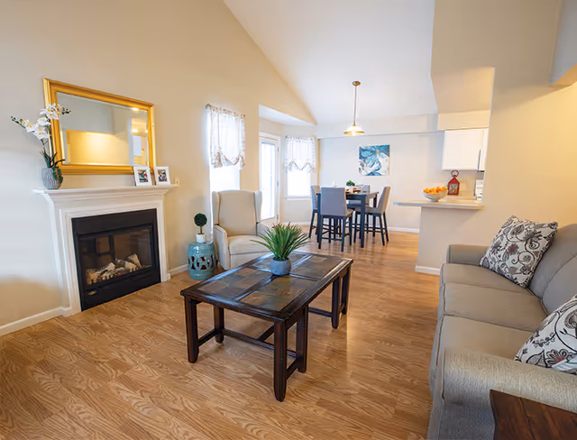 A bright and cozy living room area with a beige sofa adorned with patterned pillows, a wooden coffee table with a small potted plant, and a beige armchair next to a decorative blue ceramic stool. There is a white fireplace with a gold-framed mirror and small picture frames on the mantel. In the background, a dining area with a table and four chairs is visible, along with a kitchen counter with a bowl of oranges and a red lantern. The room has wooden flooring and light-colored walls with two windows covered by sheer curtains.