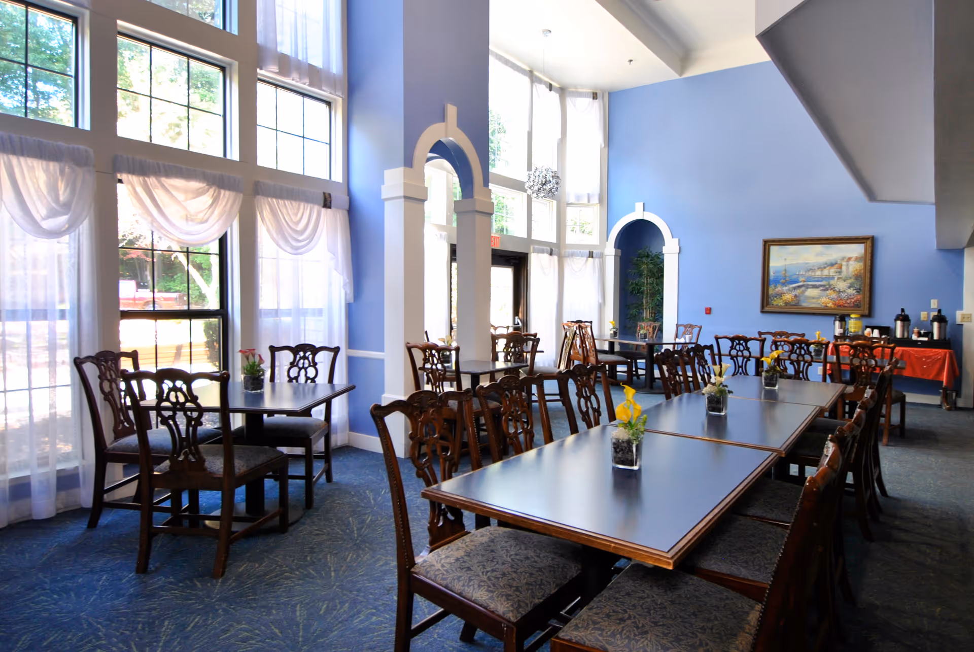 A bright dining room with large windows covered by sheer white curtains, blue walls, and multiple wooden tables and chairs arranged neatly. Small flower arrangements are placed on the tables. There is a painting on the wall and a refreshment station with coffee dispensers in the back corner.