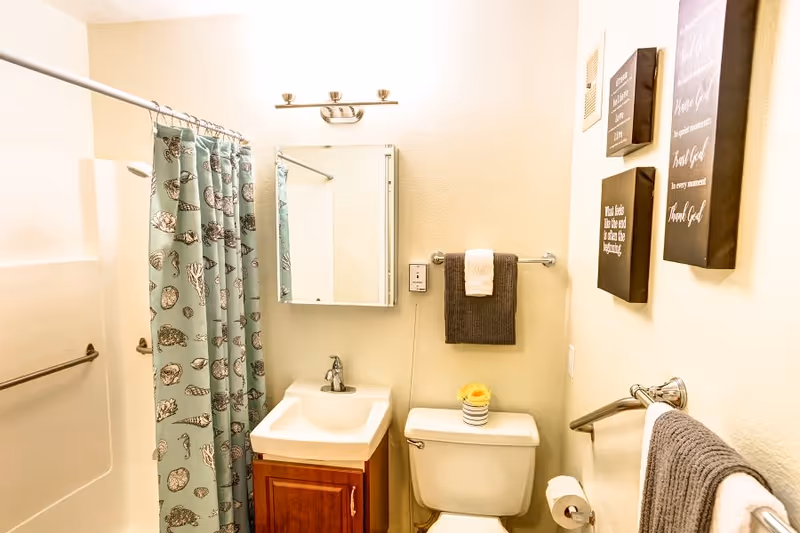A small bathroom featuring a shower with a blue patterned curtain, a white sink with a wooden cabinet underneath, a mirror with a light fixture above it, a toilet with a small decorative flower on top, and several towels hanging on racks. The walls have framed inspirational quotes.