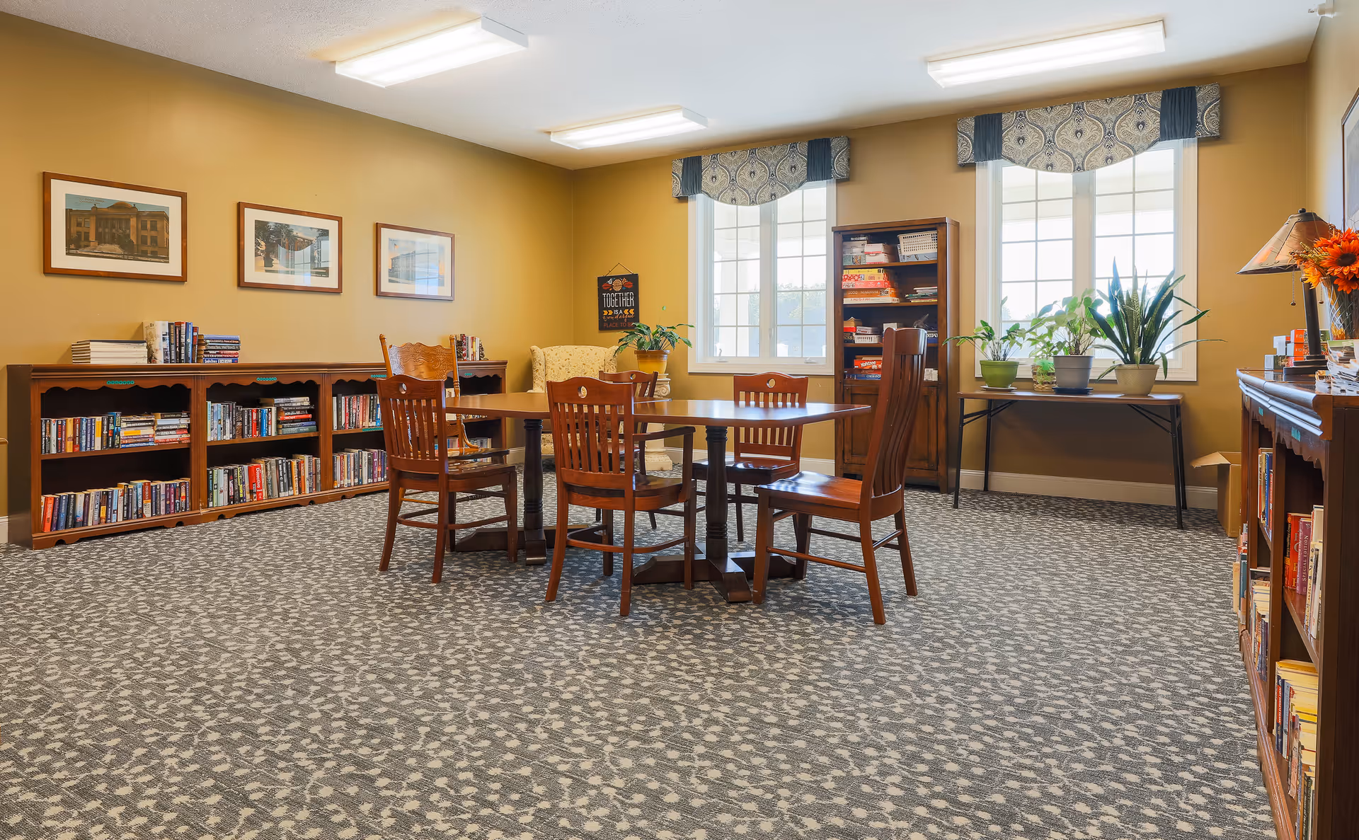A cozy reading and activity room with a round wooden table surrounded by five wooden chairs. The room has bookshelves filled with books along the walls, framed pictures, and large windows with patterned valances letting in natural light. There are several potted plants on a table near the windows and a comfortable armchair in the corner.