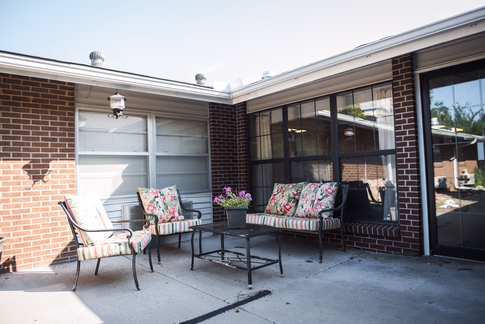 Outdoor patio area with brick walls, featuring a seating arrangement of two cushioned chairs and a cushioned loveseat with floral and striped patterns around a black metal table with a flower pot on top.