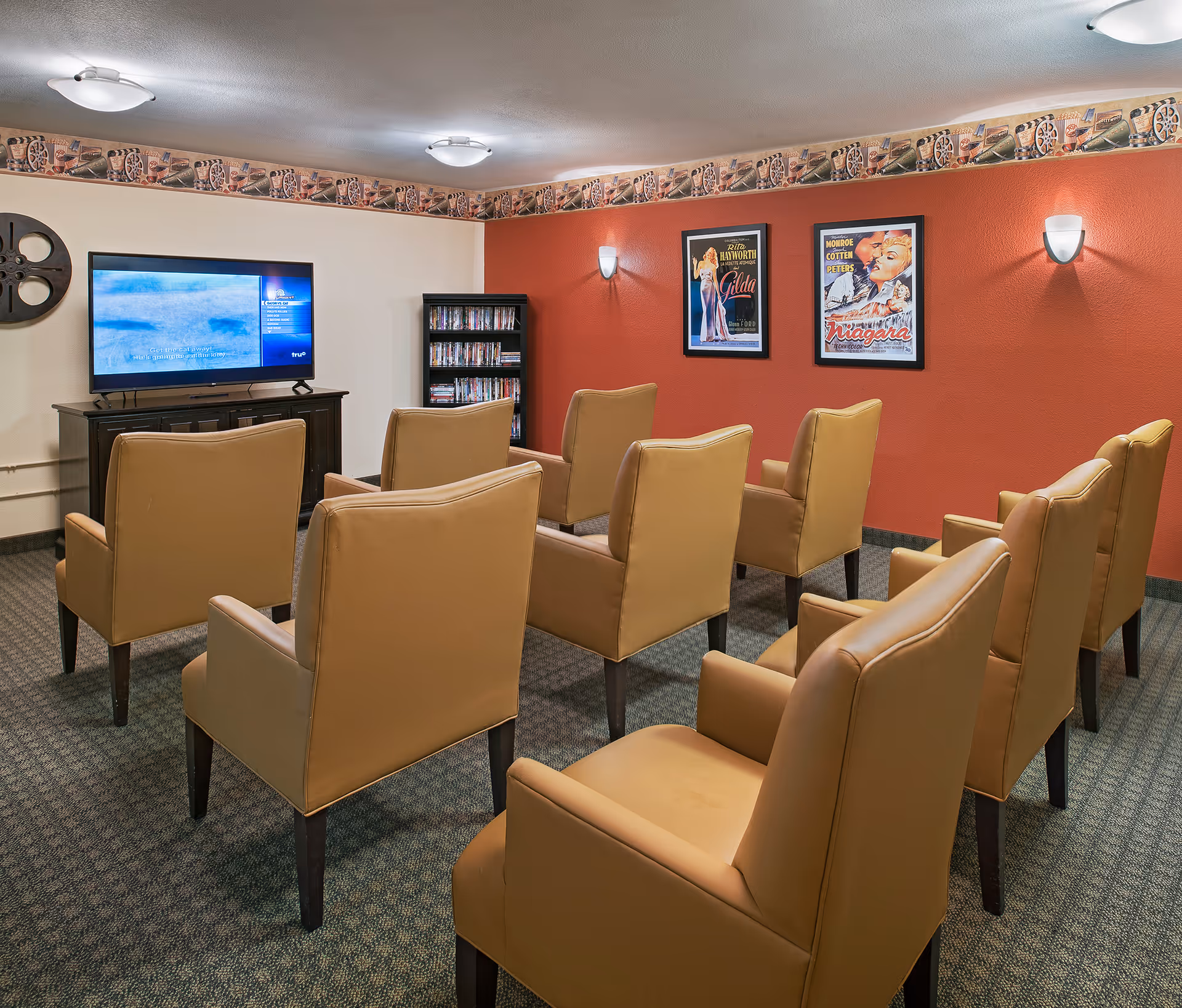A small movie theater room with eight tan leather chairs arranged in two rows facing a flat-screen TV mounted on a dark wooden stand. The walls are painted white and red, decorated with two framed vintage movie posters and a film reel wall decoration. A small bookshelf with DVDs is placed in the corner.