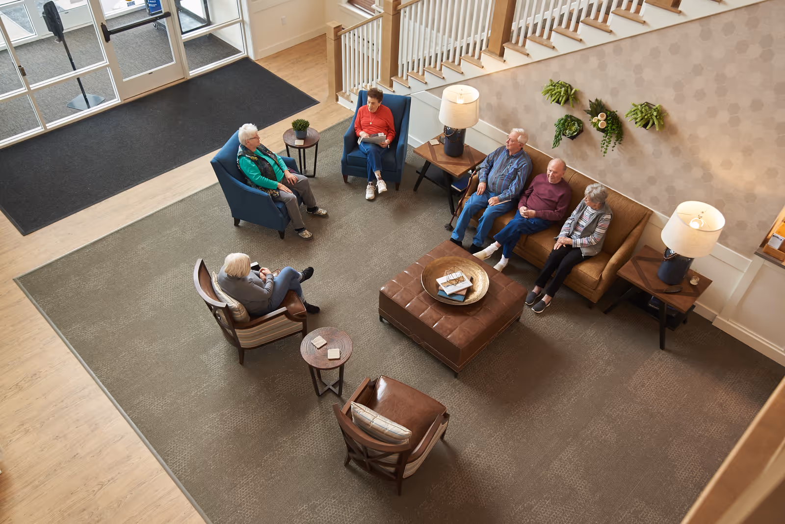 Overhead view of a senior living community lounge with residents seated around chairs and a central ottoman near an entrance and staircase.