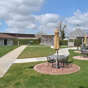 Sunny courtyard with patio tables and umbrellas, a gazebo, walkways, and surrounding single-story buildings under a partly cloudy sky.