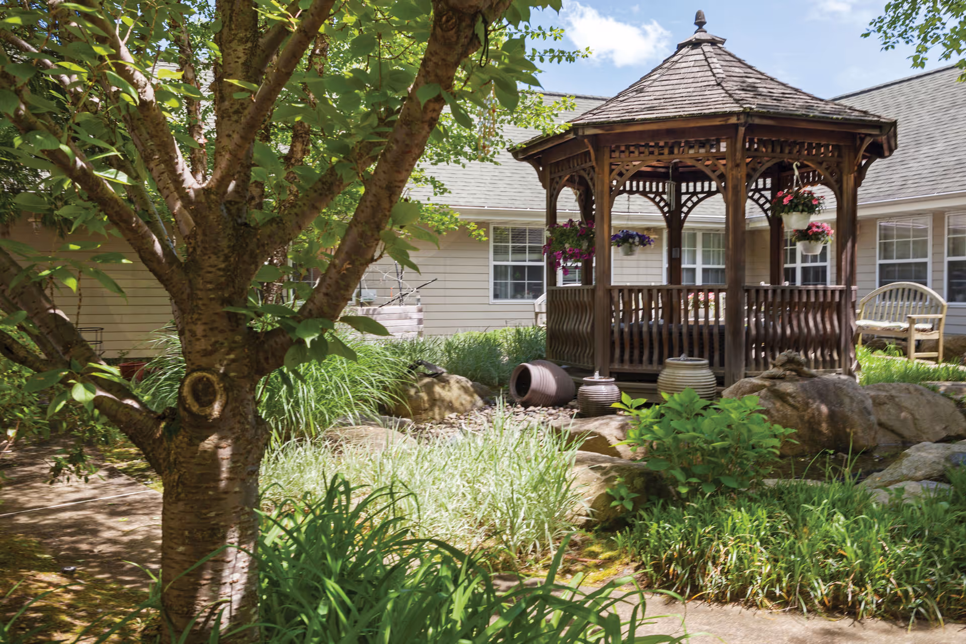 A peaceful outdoor garden area featuring a wooden gazebo with hanging flower pots, surrounded by green plants, rocks, and a tree in the foreground. The background shows a beige building with multiple windows and a bench.