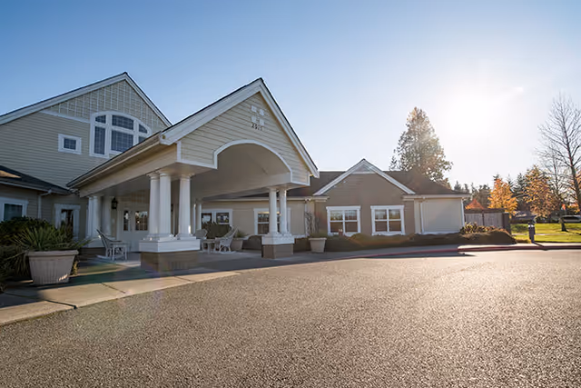 Exterior view of Brookdale Silver Lake facility entrance with a covered driveway supported by white columns, beige siding, and large windows. The sun is shining brightly in the clear sky, casting light on the paved driveway and surrounding landscaping with trees and shrubs.