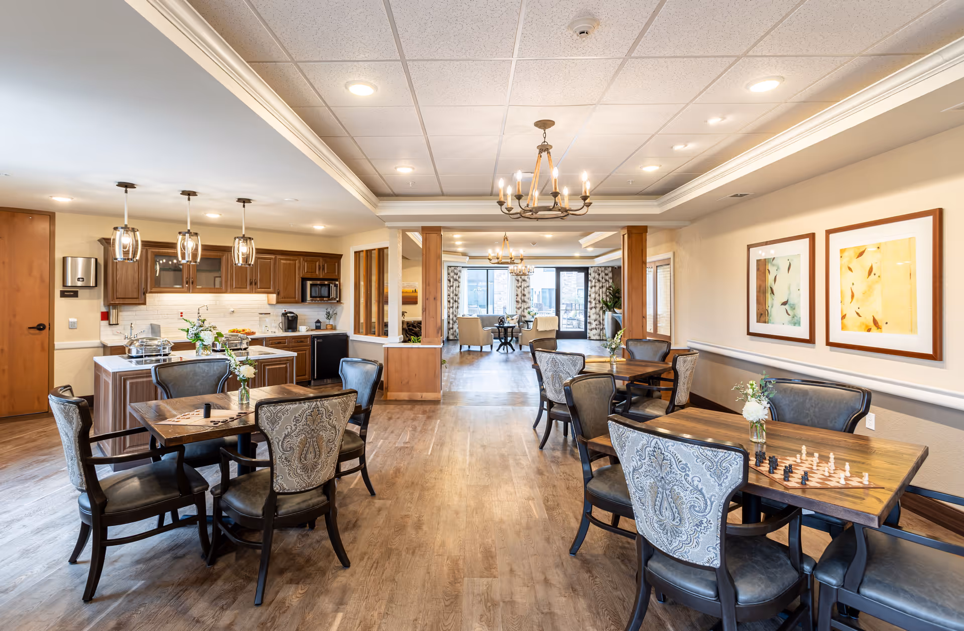 Bright communal dining and activity room with multiple tables and chairs, a kitchenette, chandeliers, and a seating area in the back.