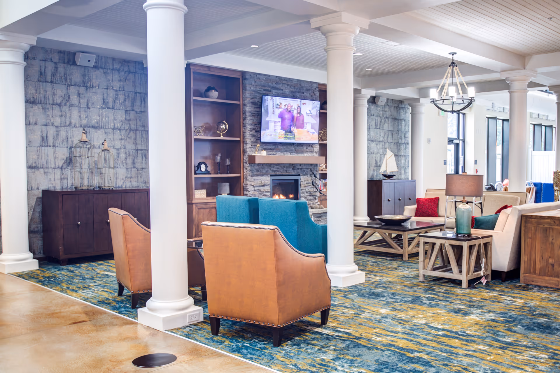 A spacious and well-lit living room area with white columns, a blue and yellow patterned carpet, and various seating options including tan leather chairs and beige sofas with colorful cushions. A stone fireplace with a mounted TV above it is centered on the far wall, flanked by built-in wooden shelves. The room features modern lighting fixtures and large windows allowing natural light to enter.