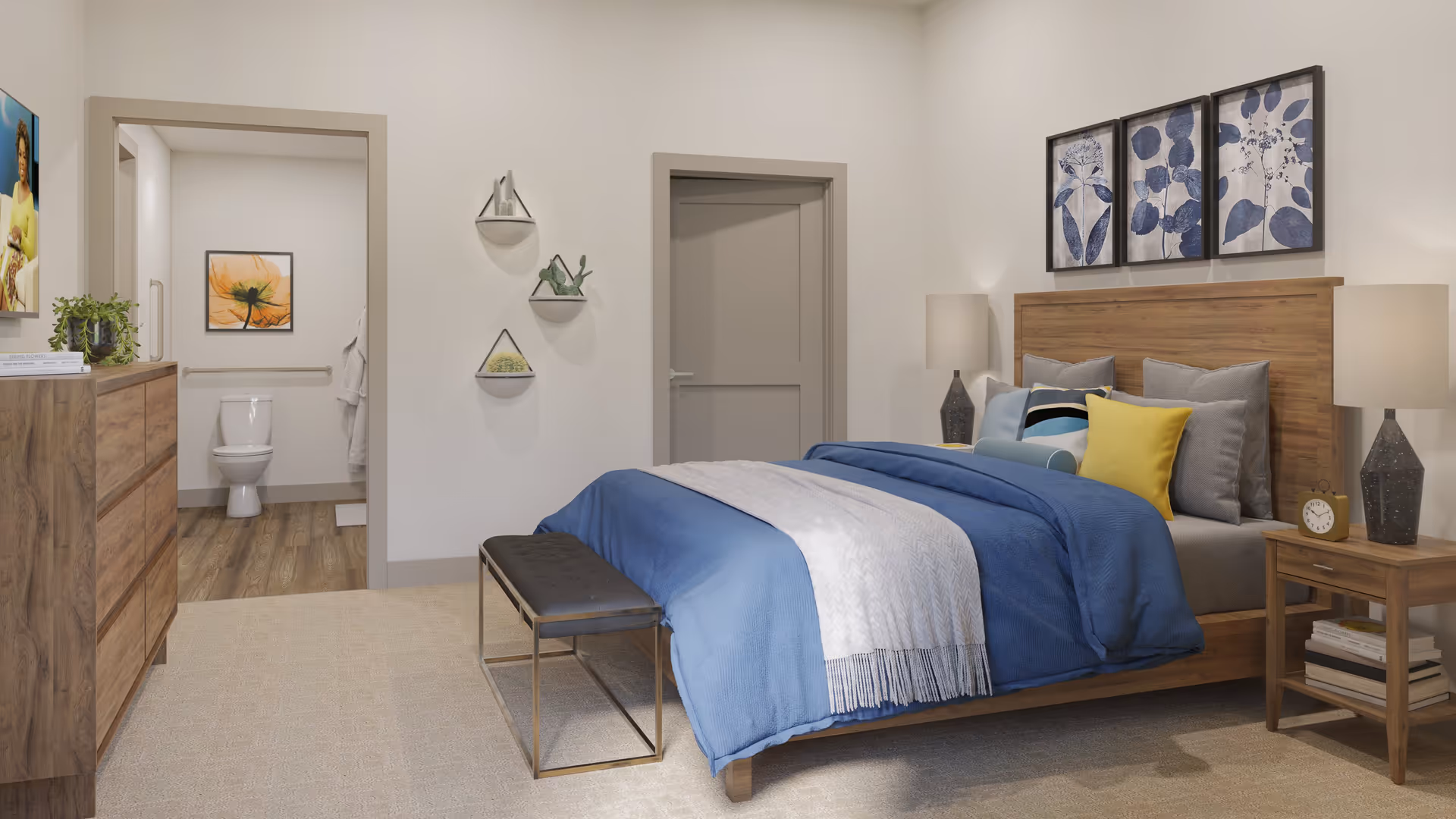 A cozy bedroom with a wooden bed frame, blue and gray bedding, and multiple pillows including a yellow one. There are two bedside tables with lamps and books, a bench at the foot of the bed, and a wooden dresser with a plant and books on top. On the wall above the bed are three framed botanical prints. To the left, an open door reveals a bathroom with a toilet, a grab bar, and a framed flower picture on the wall. The room has light-colored walls and carpeted flooring.
