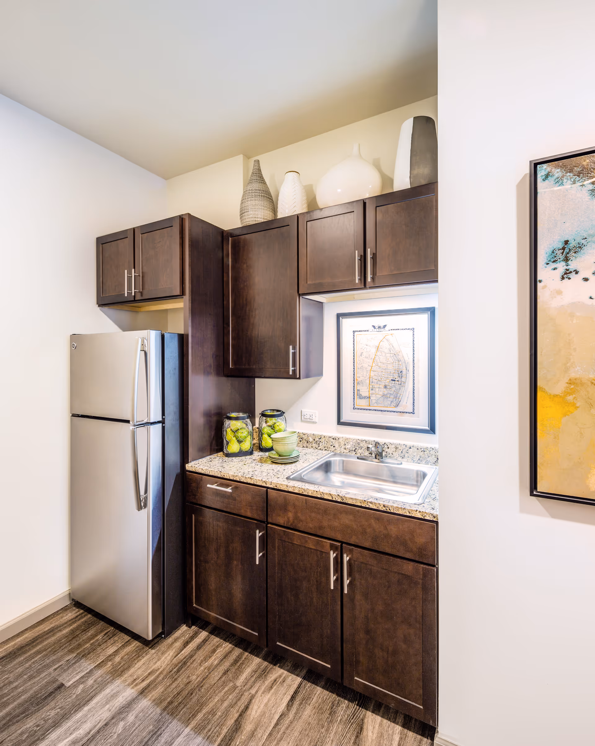 A small kitchen area with dark wood cabinets, a stainless steel refrigerator, a granite countertop with a built-in sink, two glass jars filled with green apples, stacked green bowls and plates, and decorative vases on top of the cabinets. A framed picture is hung on the wall above the sink.