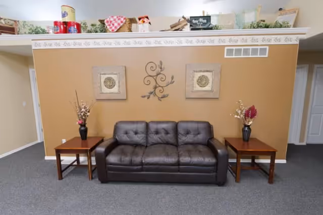 A cozy seating area with a dark brown leather sofa centered against a tan wall. On either side of the sofa are wooden side tables with decorative vases holding dried floral arrangements. Above the sofa, there are two framed square artworks and a metal wall decoration with a leaf design. The wall has a decorative border near the ceiling with the words 'LOVE LAUGH' repeated. The floor is carpeted in gray, and the room has a warm, inviting atmosphere.