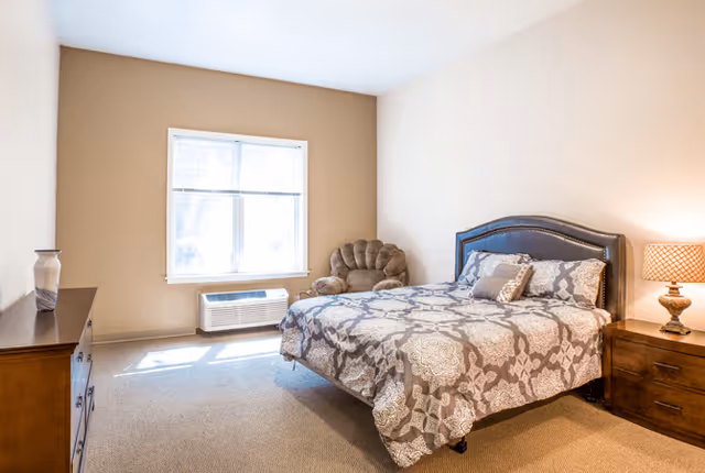 A bright bedroom with a large window letting in natural light. The room features a bed with patterned bedding, a cushioned armchair in the corner, a wooden dresser with a decorative vase, and a wooden nightstand with a lamp.