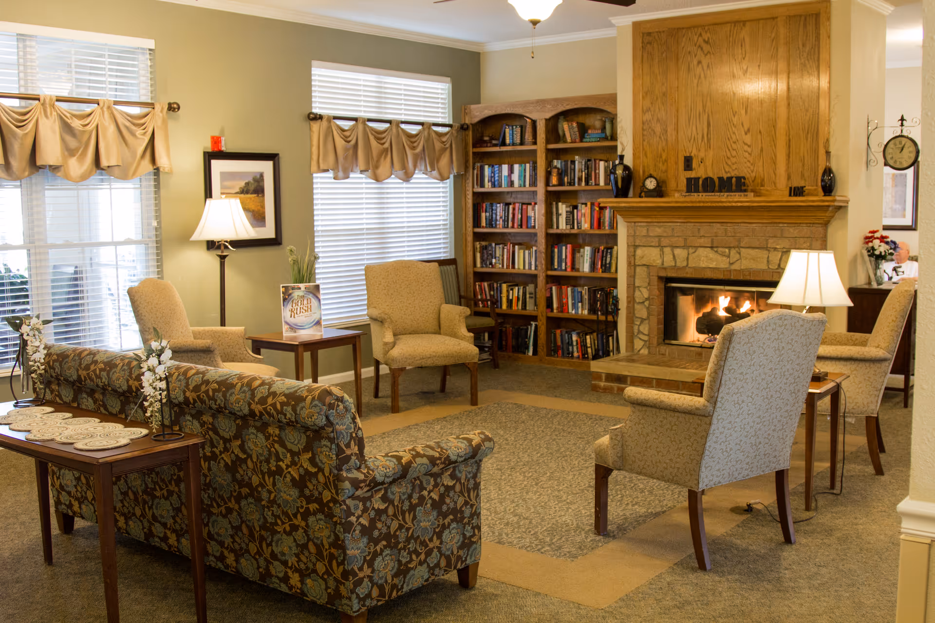 A cozy living room area with a floral patterned sofa, four beige armchairs, two side tables with lamps, a fireplace with a wooden mantle, and a bookshelf filled with books. The room has two windows with beige valances and a carpeted floor.