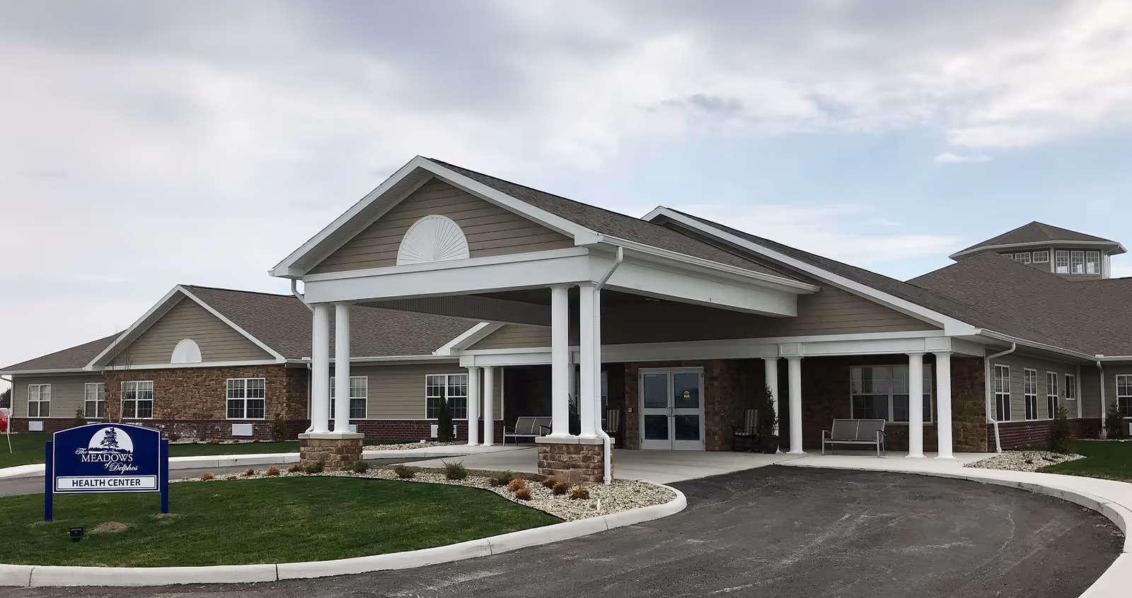 Exterior view of The Meadows of Delphos Health Center building with a covered entrance supported by white columns, surrounded by a paved driveway and landscaped grass areas under a cloudy sky.