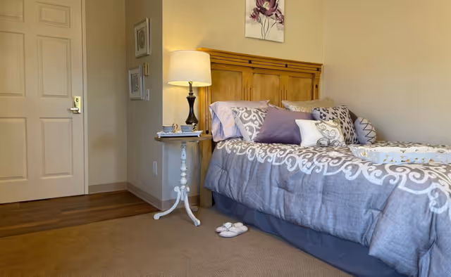 A cozy bedroom with a wooden headboard bed dressed in a gray and white patterned comforter and multiple pillows in shades of purple and white. A small white side table with a lamp, a cup, and a book is next to the bed. A pair of slippers is placed on the carpeted floor beside the bed. The room has beige walls, a closed door, and framed artwork on the wall.