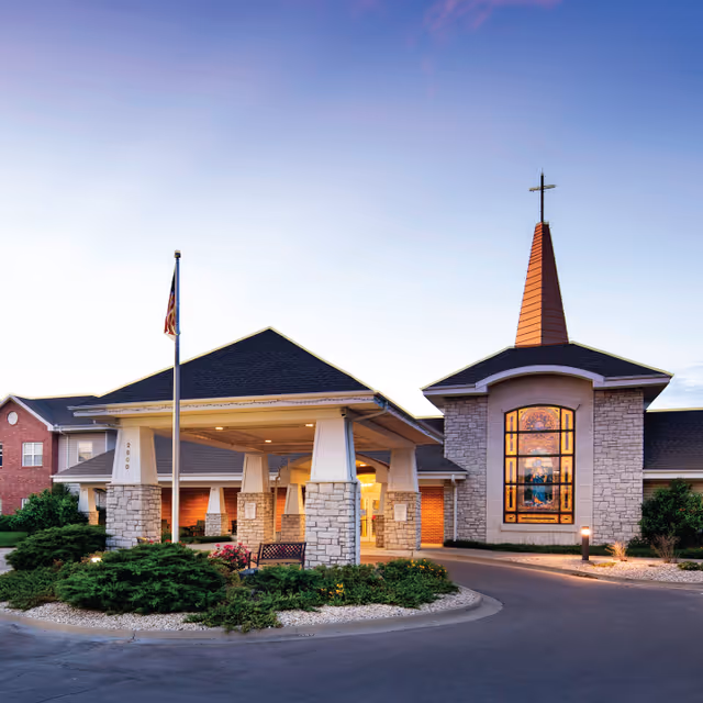 Exterior view of Ascension Living Via Christi Village building at dusk, featuring a covered entrance supported by stone pillars, a flagpole with an American flag, and a tall steeple with a cross on top. The building has brick and stone walls with a large stained glass window illuminated from inside.