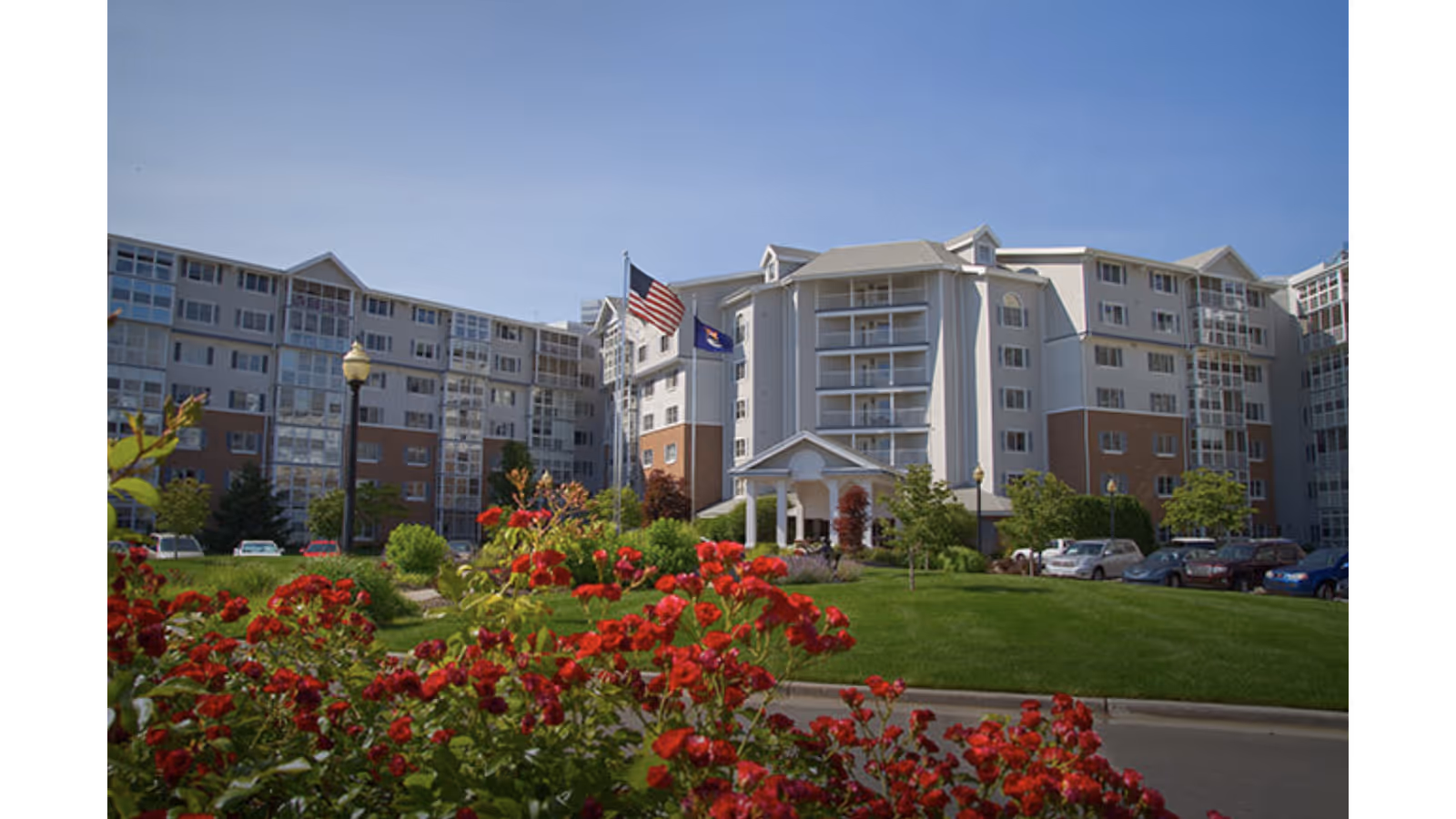Exterior view of a multi-story senior living facility with a well-maintained lawn and vibrant red flowers in the foreground. The building has multiple balconies and large windows, with two flagpoles displaying the American flag and another flag near the entrance.