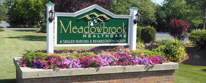 Outdoor sign for Meadowbrook Healthcare, a skilled nursing and rehabilitation facility, surrounded by a flower bed with purple and pink flowers and green shrubs in a landscaped area.