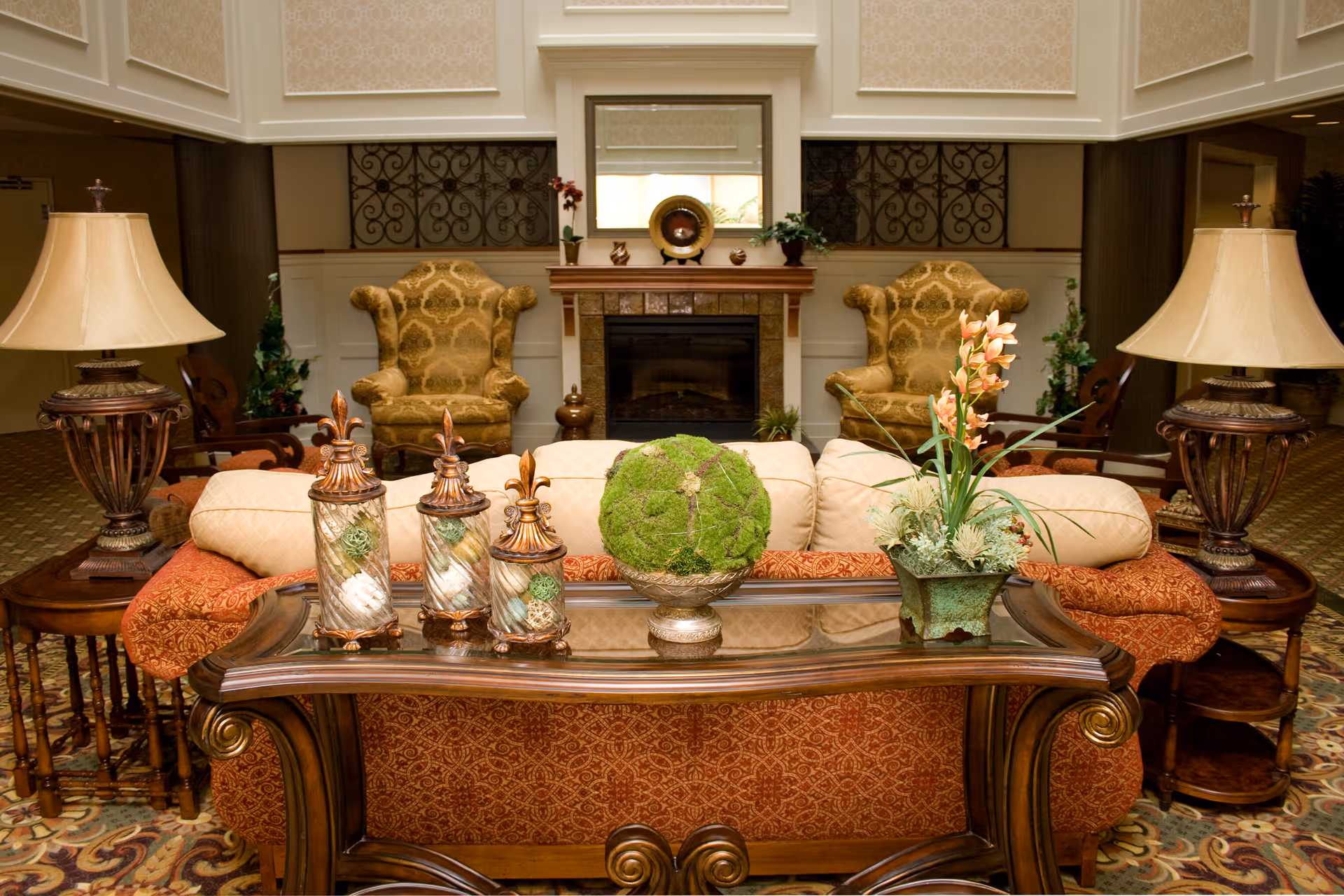 A cozy living room area with a patterned orange sofa, two ornate armchairs, a glass-top wooden coffee table with decorative items including a moss ball and a flower arrangement, two side tables with lamps, and a fireplace with a mirror above it in the background.