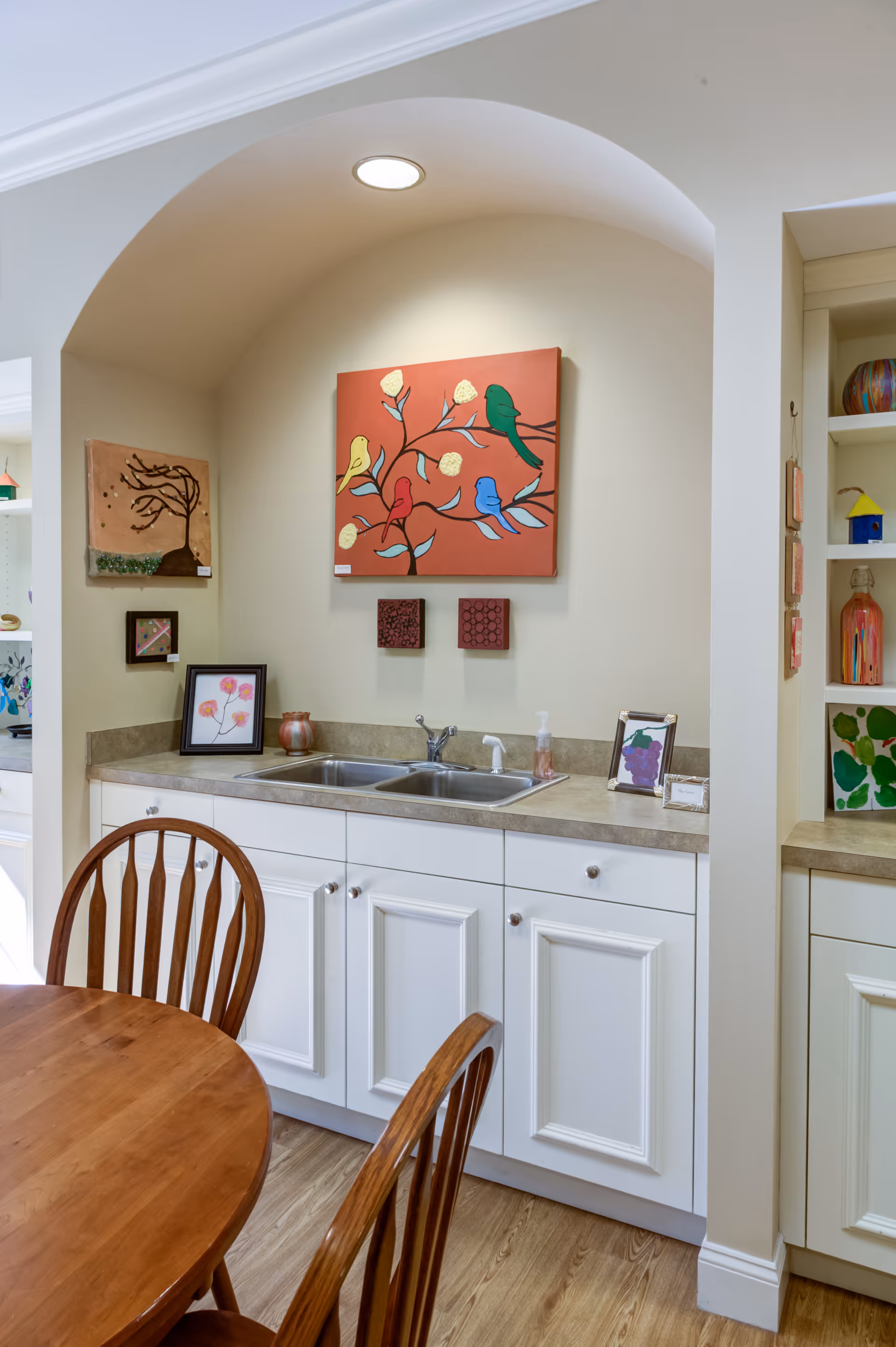 Interior view of a kitchen area with a double sink set in a beige countertop and white cabinets below. Above the sink, there is a colorful painting of birds on a branch mounted on the wall under a recessed light. To the left, there is a wooden dining table with matching chairs. Shelves with decorative items are visible on the right side.