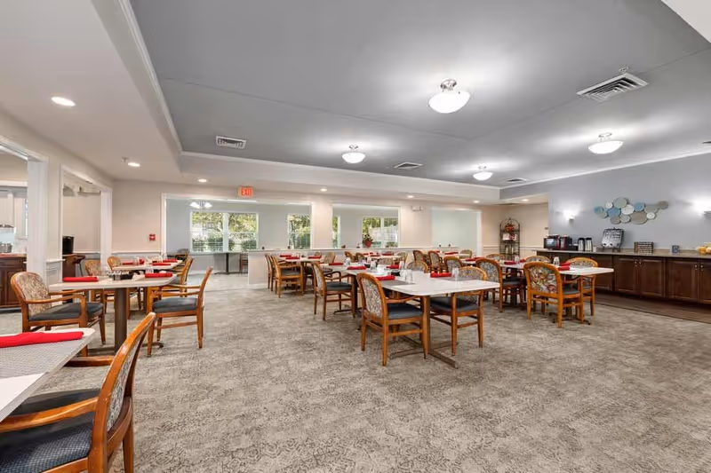 A spacious dining room with multiple tables and chairs arranged neatly. Each table has red napkins and glassware. The room has large windows allowing natural light to enter, beige walls, and a carpeted floor. There is a counter area with coffee machines and decorative items on the right side of the room.