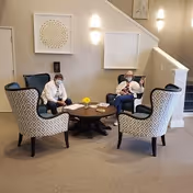 Two people wearing masks sitting in a lounge area with four patterned armchairs arranged around a round wooden coffee table with a small yellow flower centerpiece. The background shows beige walls with two framed decorative artworks and wall-mounted lights, next to a staircase.