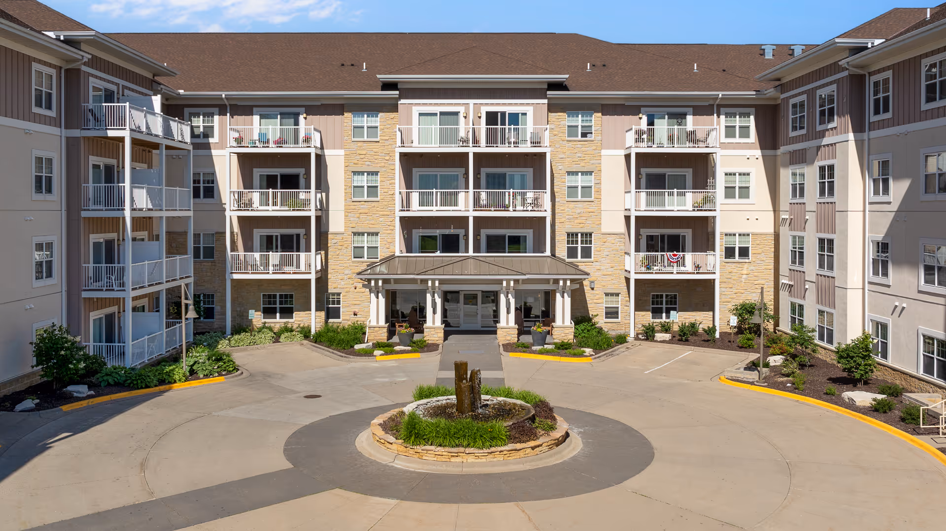 Front exterior view of McKenna Crossing, a multi-story senior living building with balconies, a covered entrance, and a circular driveway with a landscaped center island featuring a small water fountain.