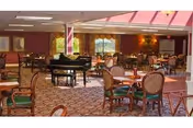 Spacious dining room with multiple round tables and chairs arranged neatly. A black grand piano is positioned near the center of the room. The room features large windows with curtains, patterned carpet flooring, and warm lighting from ceiling fixtures.